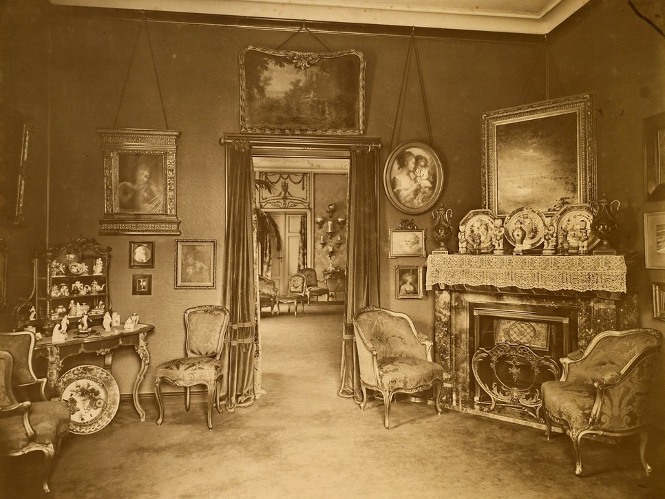 Historische Sepiafotografie eines Zimmers in der Wiener Straße 25. an den Wänden hängen verschiedene Gemälde auf einem Kamin und einem kleinen Schränkchen stehen verschiedene Porzellane.