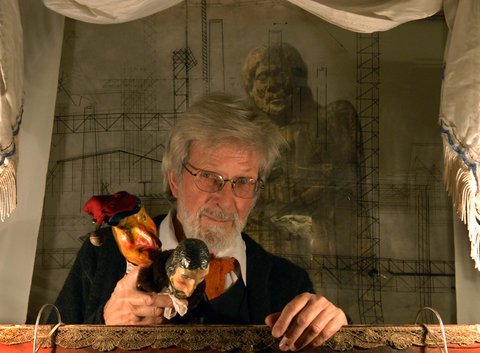 A bearded man smirks into the camera. He is standing inide a stage construction and holding two puppets.