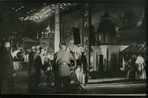 Ein Foto von den Dreharbeiten an Prometheus. Im Hintergrund ist die Filmkulisse zu sehen, die wie auf einer Theaterbühne mit Gebäuden bestellt und einem großen Gemälde hinterlegt ist. Oben sind die Schweinwerfer zu sehen im Vordergrund stehen zwei kostümierte Schauspieler und die Kameraleute.