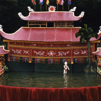 Wasserpuppentheater im Großen garten. Die Bühne besteht aus einem großen Wasserbassin, über dem die Puppen bewegt werden, den Hintergrund des Bühnenbilds bildet ein Haus mit Pagodendach.