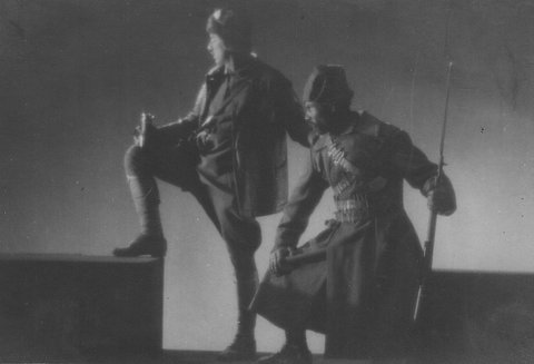 Filmstill in Schwarzweiß, das zwei Soldaten in Profilansicht zeigt. Der eine sitzt, der andere steht und hat ein Bein auf ein Podes (einer der grauen Blöcke im Text) gestellt, was ihm eine kühne Haltung verleiht. In der rechten hand hält er einen Becher. Sie sind nach links gewandt und schauen in die Ferne.