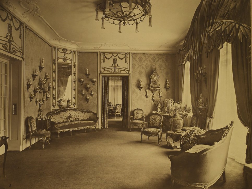 Historische Sepiafotografie des Salons in der Wiener Straße 25. An den Wänden stehen Porzellane auf kleinen Wandpodesten.