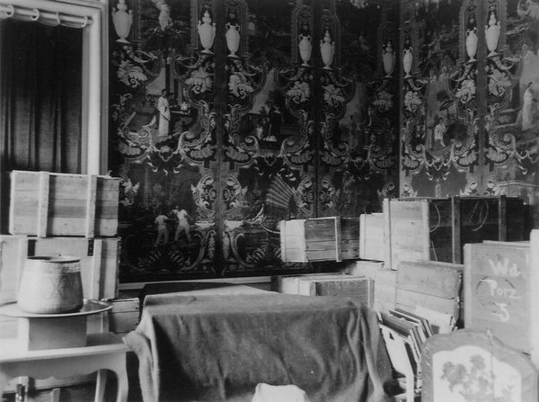 Schwarz-weißfotografie. Innenraumaufnahme mit gestapelten Holzkisten. Am den Wänden ist eine prunkvolle Tapete angebracht.