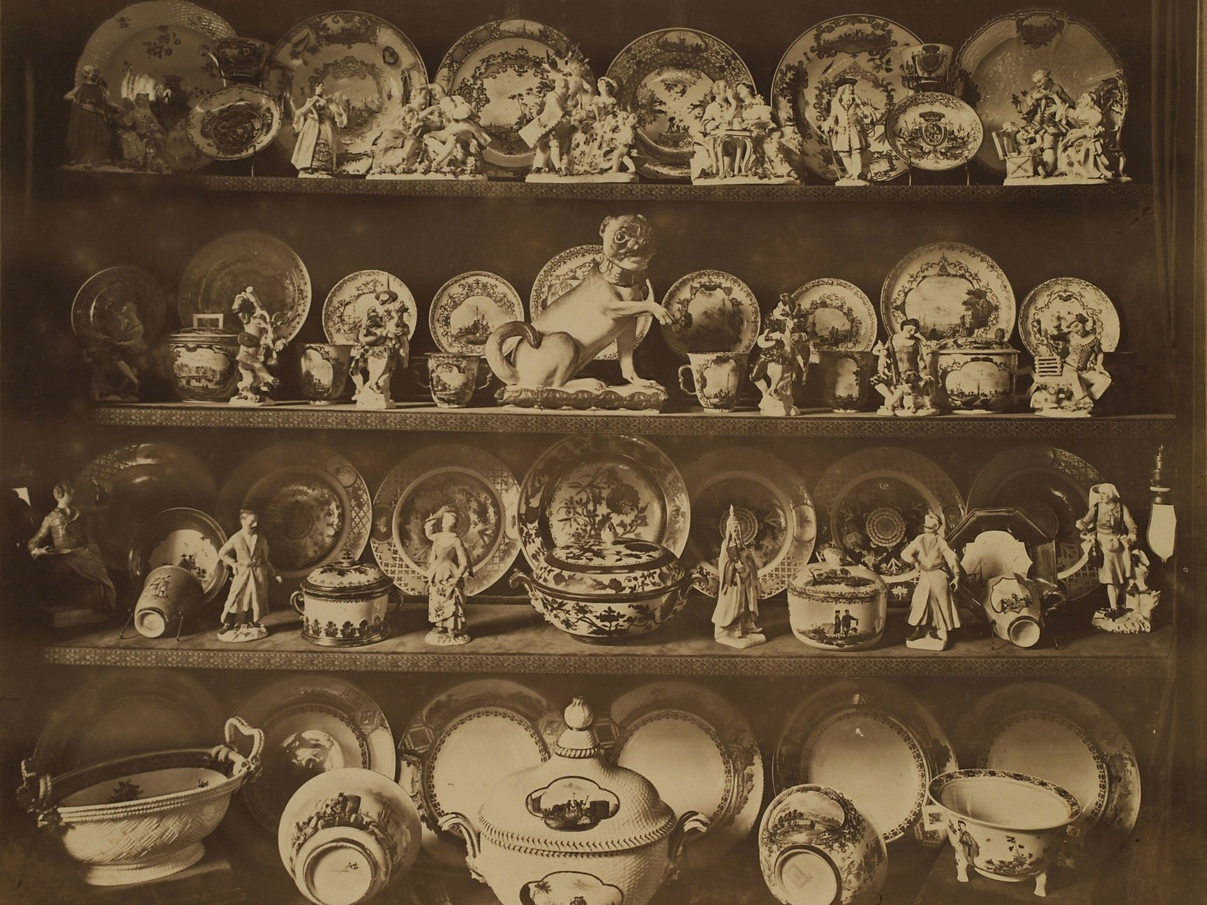 Historische Sepiafotografie. Auf drei Regalbrettern übereinander stehen Porzellanfiguren und Teller. In der Mitte befindet sich ein Porzellanmops.