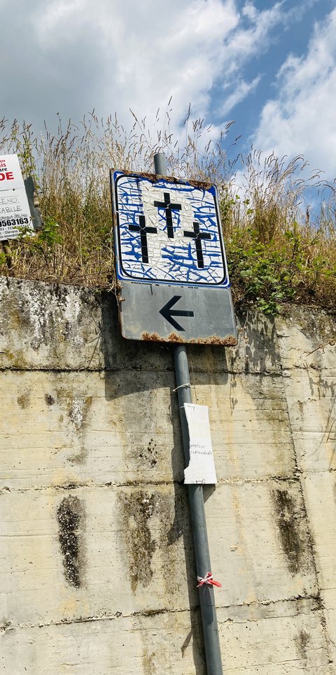Ein Schild, das mit drei Kreuzen auf eine Gedenkstätte hinweist und das selbst starke Verschleißspuren zeigt. Die alte blaue Farbe ist so rissig, dass sie die Fläche um die drei Kreuze nur noch fetzenhaft bedeckt.