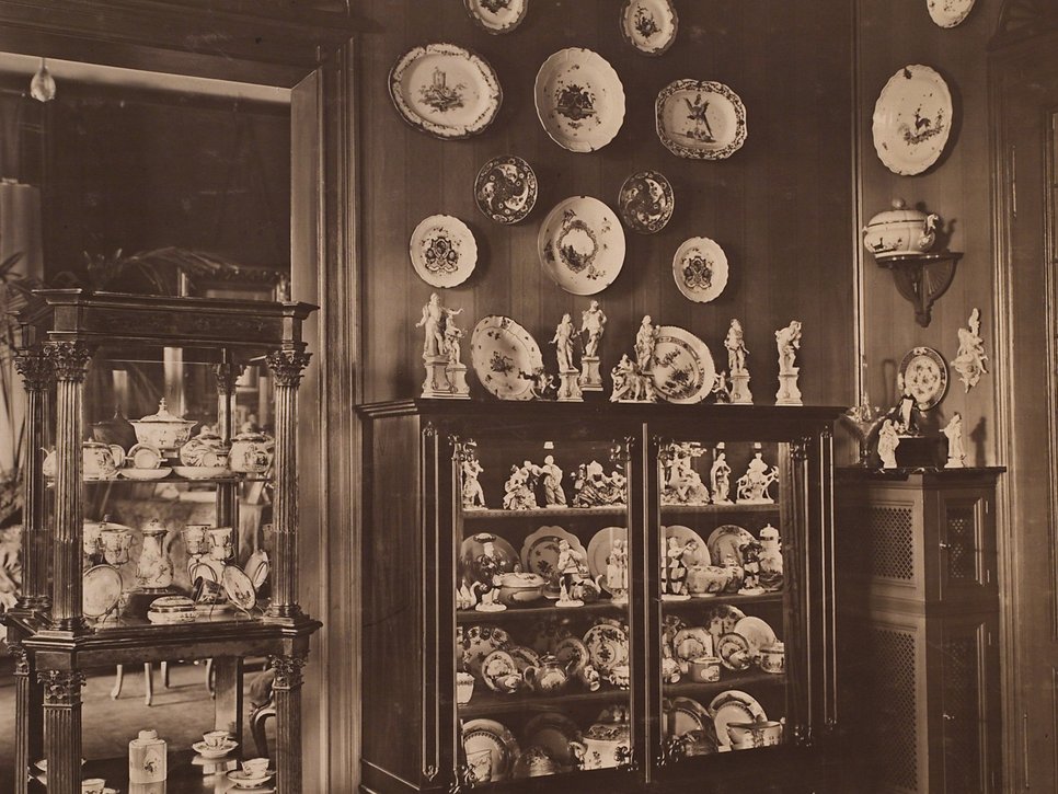 Sepia Fotografie, historische Aufnahme eines Innenraumes. Eine hohe, mit Porzellan gefüllte, Holzvitrine steht zur rechten Seite einer Türöffnung. In ihrer Mitte befindet sich eine weitere gefüllte Vitrine. Um den Türrahmen sind dekorative Teller gehängt.