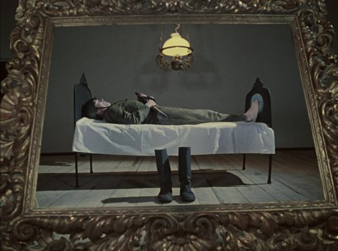 Durch einen großen goldenen Barock-Rahmen hindurch ist horizontal ein liegender Soldat in einem Bett liegend zu sehen. Er ist uniformiert und hält sein Gewehr über der Brust. Vor dem Bett stehen seine Stiefel, über ihm hängt mittig eine Lampe.