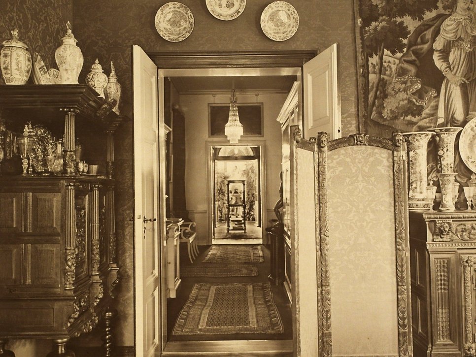 Sepia Fotografie, historische Aufnahme eines Innenraumes in der Wiener Straße 25. Der Blick geht durch eine offene Tür einen langen Gang entlang. Um den Türrahmen sind dekorative Teller gehängt.