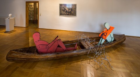 Die Ausstellungsräume in Glauchau: In einem Holzboot sitzen zwei Figuren, die eine hält ein Handy, die andere trägt eine Schwimmweste. Zwischen ihnen ist das Boot in der Mitte mit Stacheldraht überwölbt. An der Wand im Hintergrund hängt ein Gemälde von Anja Elze.