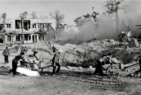 Eine alte Fotografie des zerstörten Vinh. Im Vordergrund ziehen einige Personen einen Karren durch den Schlamm, ahinter sind Schuttberge und zerstörte Häuser zu sehen.