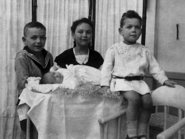 Schwarz-weiß Aufnahme der vier Kinder von Victor und Sophie von Klemperer. Auf einem Tisch liegt ein Neugeborenes in weißer Kleidung. Hinter dem Tisch stehen ein Junge und ein Mädchen. Auf der rechten Kante des Tisches sitzt ein kleiner Junge.