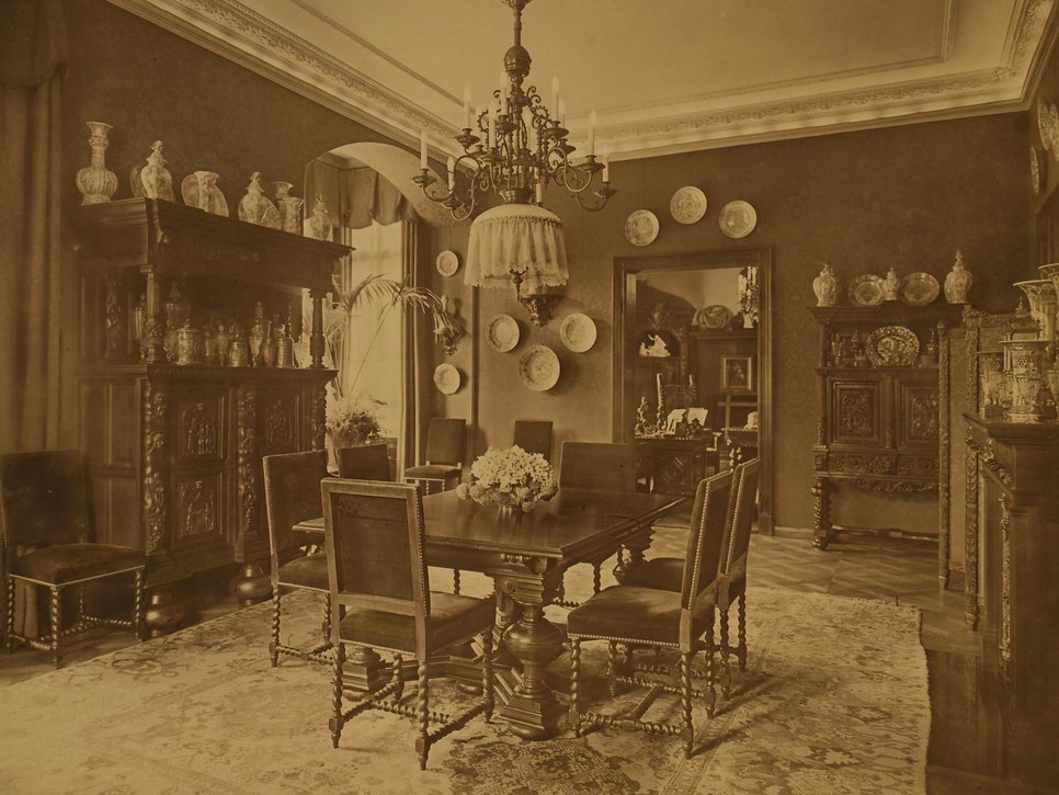 Historische Sepiafotografie eines Zimmers in der Wiener Straße 25. Auf historischen Holzmöbeln steht eine Vielzahl von Porzellanvasen. In der Mitte des Raums befindet sich ein Holztisch.