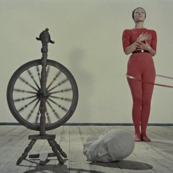 Links ein Spinnrad, rechts im Hintergrund eine Frau in einem engen roten Kostüm mit Hula-Hoop-Reifen. Dazwischen liegt der steinerne Kopf einer Skulptur.