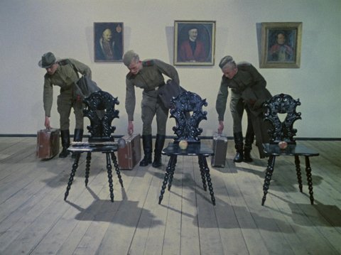 Drei Soldaten in Frontalansicht stellen synchron ihre Koffer ab. Im Verdergrund stehen drei Stühle an der Wand hinter ihnen hängen drei Portraits von Adeligen.