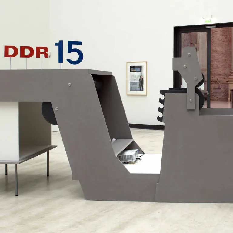 Ein grauer maschinenhafter Aufbau im Lipsiusbau. In das graue, mit einigen Schrauben besetzte Objekt sind zwei schwarze Walzen integriert, sodass es an die Maschinen einer Zeitungsdruckanlage erinnert. Das Objekt ist zugleich Rahmung und Verdunkelung für eine Beamerprojektion, die darin gesehen werden kann. Obendrauf sind an dünnen, nach oben ragenden Stelzen die Zeichen "DDR 15" angebracht, wobei die Buchstaben in sattem Rot und die Zahl in dunklem Blau gefärbt ist.
