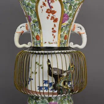 Komplettansicht der Vase: Nach oben hin ist sie trompetenartig geöffnet und farbenfroh mit floralen Elementen bemalt. In der Mitte, kurz oberhalb des Bauches, befinden sich die beiden Elefantenköpfe, deren einer den gedruckten Rüssel trägt. Der Bauch darunter ist von einem eingefassten Vogelkäfig umgeben, der das Ornament eines Vogels beinhaltet. Darunter der ebenfalls farbenfrohe Fuß. 