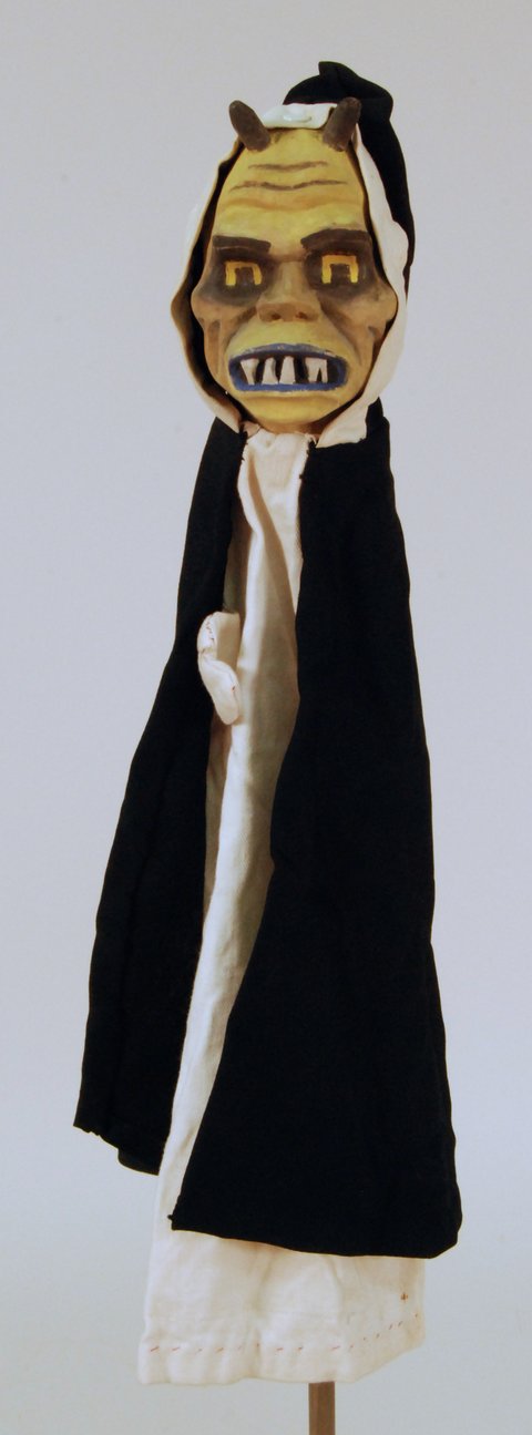 Handpuppe mit schwarzem Umhang und weißer Unterbekleidung. Das Gesicht ihat eine ungesunde Farbe, die Zähne sind spitz und die Figur hat zwei kleine Hörner, die vor der Kapuze aus dem Kopf ragen.