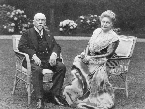 Schwarz-weiß Fotografie von Gustav und Charlotte von Klemperer. Sie sitzen in schicker Kleidung auf Korbstühlen in einem Garten und Blicken in die Kamera.