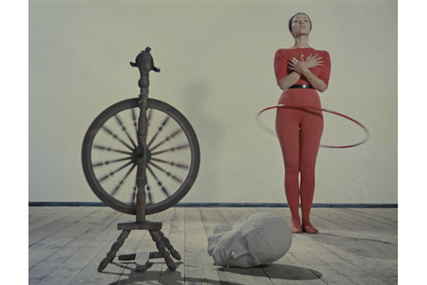 Links ein Spinnrad, rechts im Hintergrund eine Frau in einem engen roten Kostüm mit Hula-Hoop-Reifen. Dazwischen liegt der steinerne Kopf einer Skulptur.